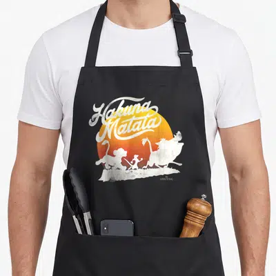 Tablier de cuisine noir avec inscription Hakuna Matata et silhouettes de personnages animés devant un soleil couchant orange et jaune.