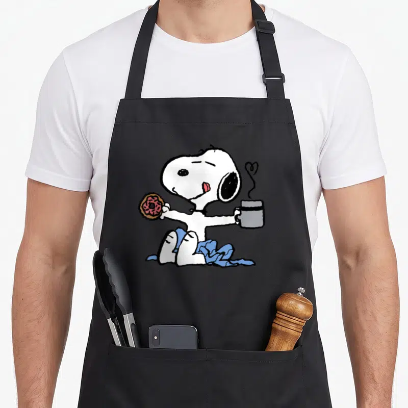 Tablier de cuisine noir avec Snopy assis tenant un donut et une tasse de café fumant.