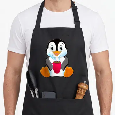 Tablier de cuisine noir avec un pingouin cartoon tenant une tasse rouge fumante et des pattes orange posées au sol.