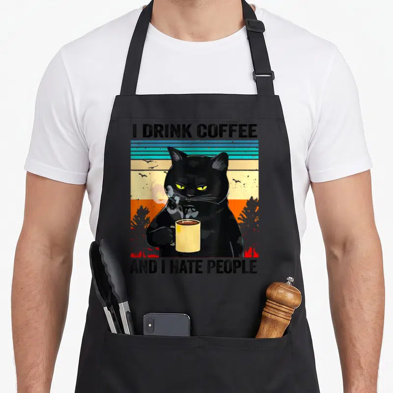 Tablier de cuisine noir avec un chat noir tenant une tasse jaune et du texte en anglais sur fond coloré.