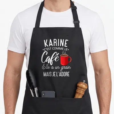 Tablier de cuisine noir avec inscription blanche Karine et illustration de tasse de café rouge fumante au centre du motif imprimé.