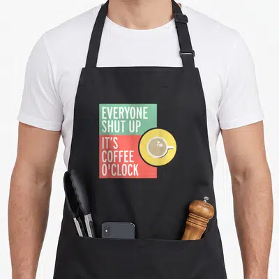 Tablier de cuisine noir avec inscription bilingue et illustration de tasse de café sur fond coloré géométrique.