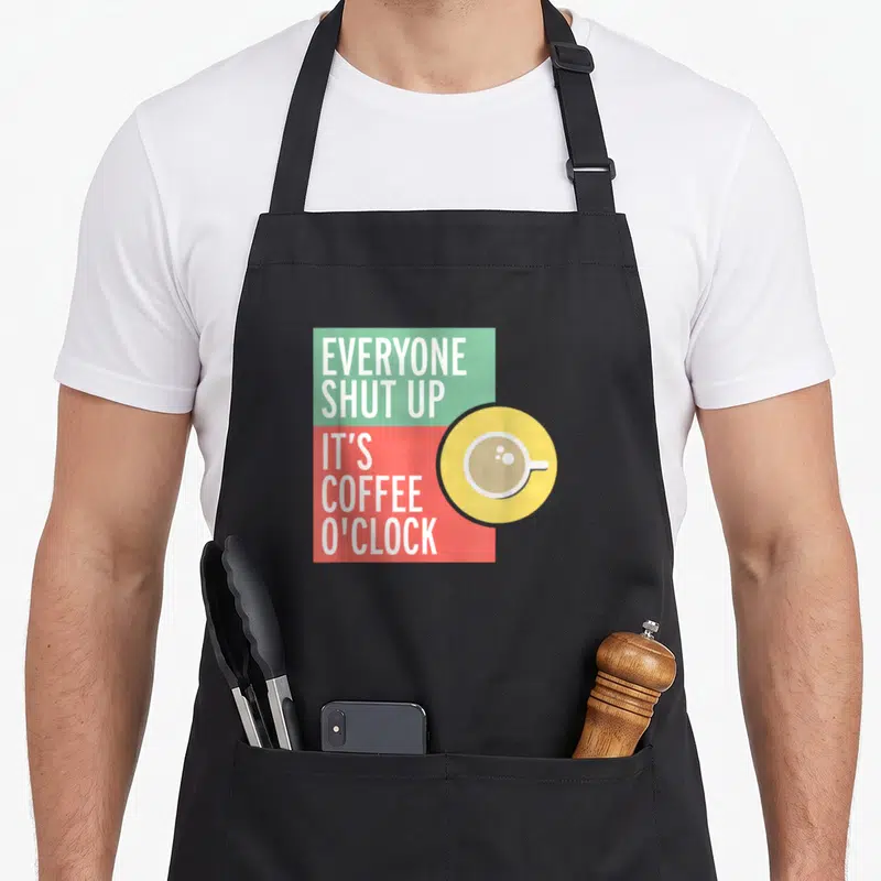 Tablier de cuisine noir avec inscription bilingue et illustration de tasse de café sur fond coloré géométrique.