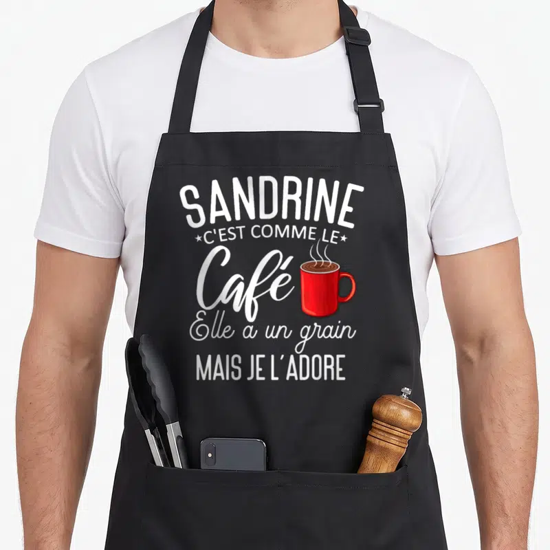 Tablier de cuisine noir avec texte blanc Sandrine et illustration d'une tasse de café rouge fumante au centre du design.
