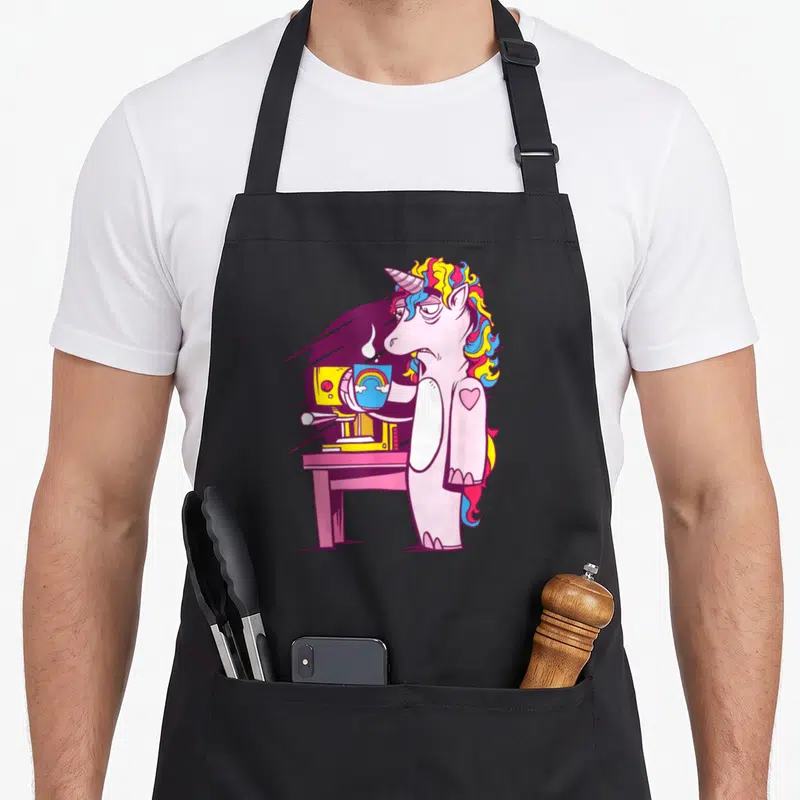 Tablier de cuisine noir avec une licorne rose épuisée tenant une tasse de café arc-en-ciel devant une machine jaune.