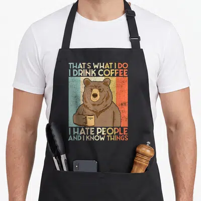 Tablier de cuisine noir avec ours brun tenant une tasse de café sur fond rayé multicolore.