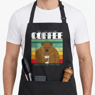 Tablier de cuisine noir avec un ours brun tenant une tasse de café sur fond de bandes colorées horizontales.