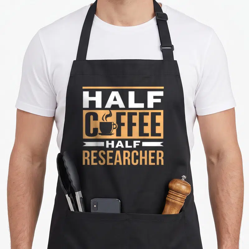 Tablier de cuisine noir avec inscription Half Coffee Half Researcher et illustration de tasse fumante orange et gris.