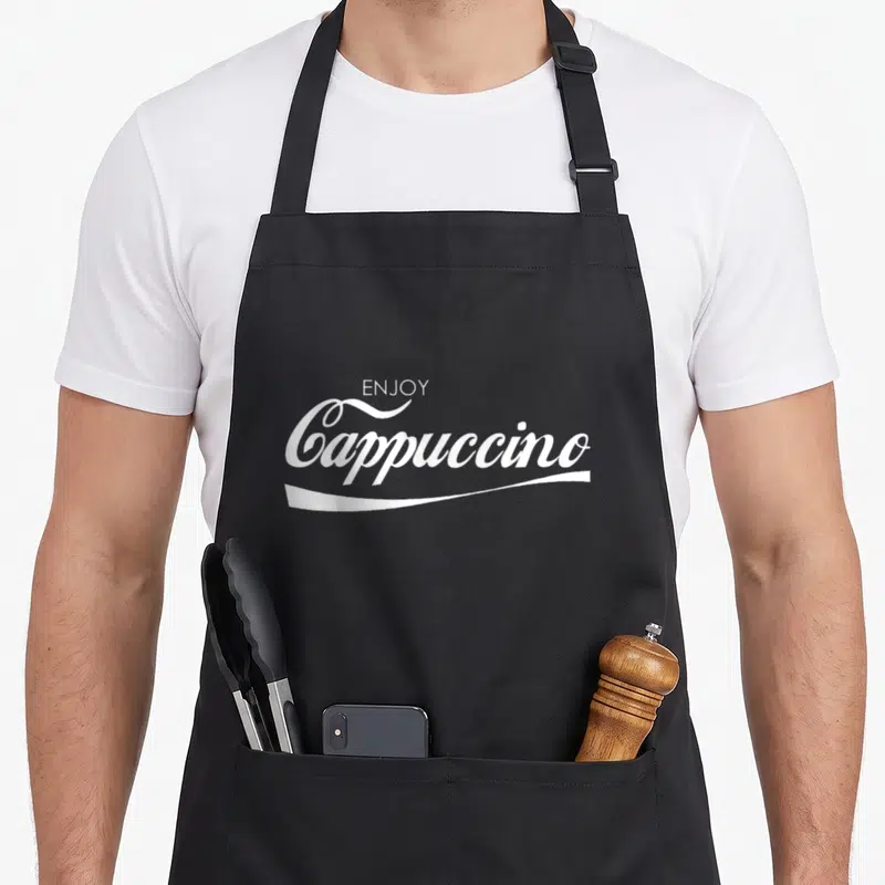 Tablier de cuisine noir avec le texte « Enjoy » Cappuccino en écriture cursive blanche stylisée.