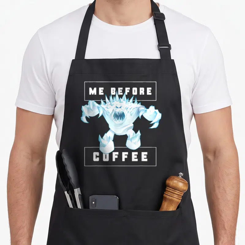 Tablier de cuisine noir avec illustration du monstre de neige et texte Me Before Coffee en blanc.