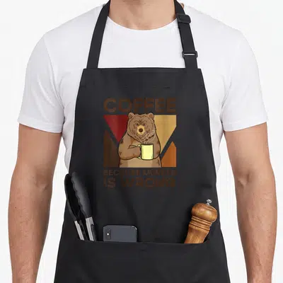 Tablier de cuisine noir avec un ours tenant une tasse jaune et le texte Coffee Because Murder Is Wrong imprimé en brun.