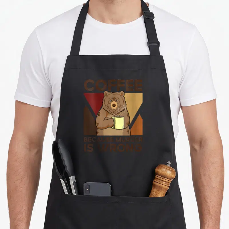 Tablier de cuisine noir avec un ours tenant une tasse jaune et le texte Coffee Because Murder Is Wrong imprimé en brun.