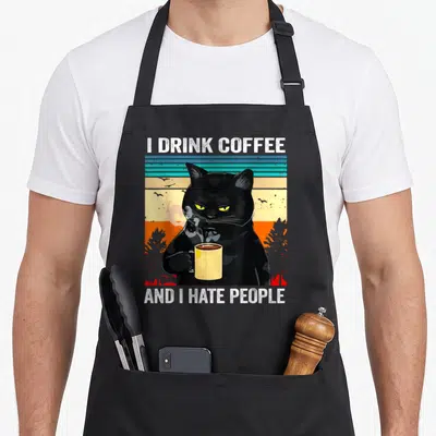 Tablier de cuisine noir avec illustration de chat noir tenant une tasse et texte en anglais.