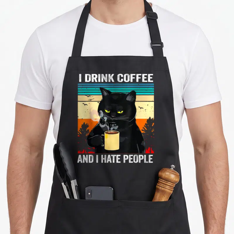 Tablier de cuisine noir avec illustration de chat noir tenant une tasse et texte en anglais.