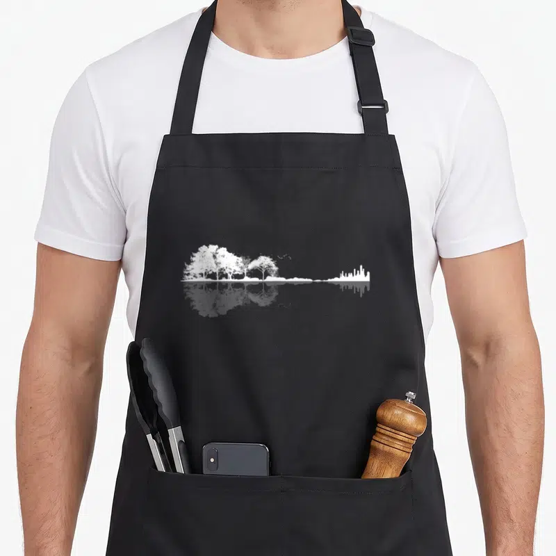 Tablier de cuisine noir avec illustration blanche de forêts et skyline urbain reflétés dans l'eau.