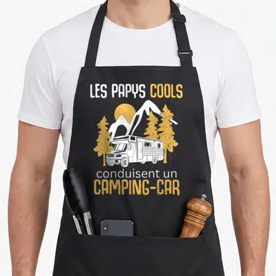 Tablier de cuisine noir avec illustration d'un camping-car blanc sous des montagnes et des arbres aux couleurs ocre et blanche.