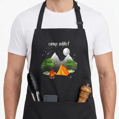 Tablier de cuisine noir avec illustration d'une tente orange, d'un feu de camp et du texte camp addict sous un ciel étoilé.