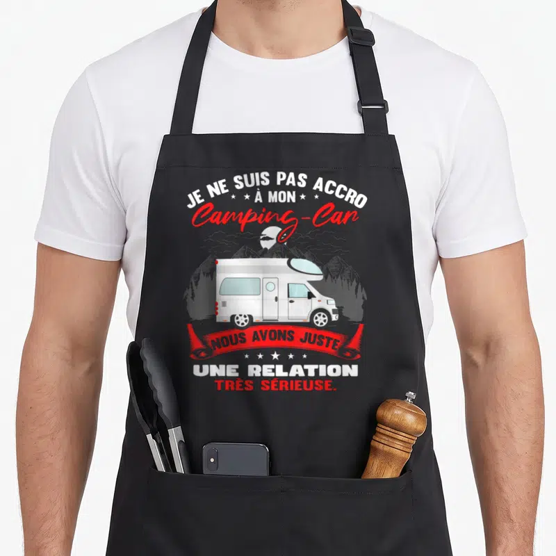 Tablier de cuisine noir avec illustration d'un camping-car blanc et texte humoristique sur fond de montagnes.