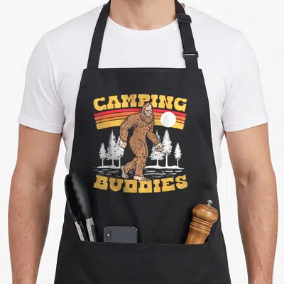 Tablier de cuisine noir avec illustration rétro d'un Bigfoot entre des sapins et le texte Camping Buddies en jaune.