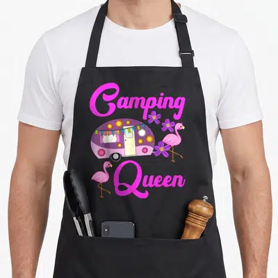 Tablier de cuisine noir avec une caravane violette fleurie, deux flamants roses et le texte Camping Queen en rose vif.