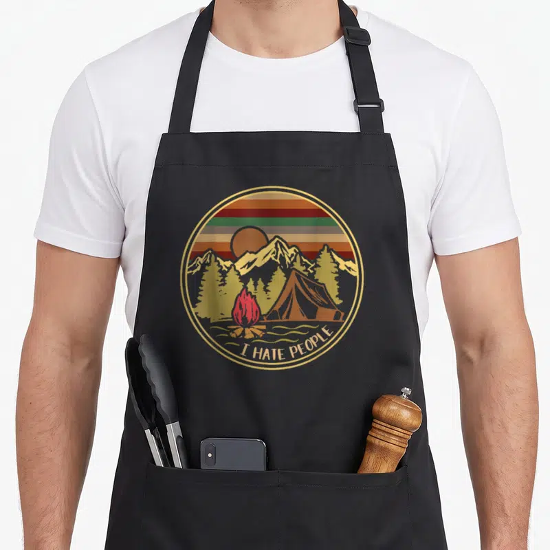 Tablier de cuisine noir avec illustration circulaire vintage représentant une tente, un feu de camp et des montagnes sous un ciel strié.