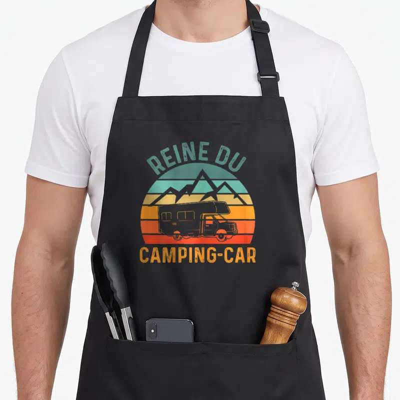 Tablier de cuisine noir avec illustration rétro d'une caravane devant des montagnes et un soleil couchant aux couleurs dégradées.