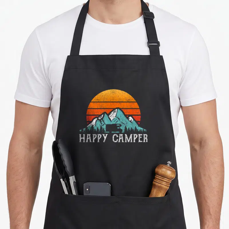 Tablier de cuisine noir avec illustration retro de soleil orange, montagnes bleues et texte Happy Camper en blanc