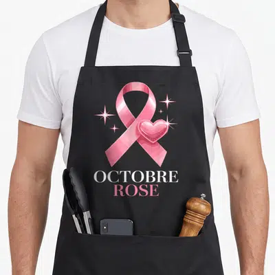 Tablier de cuisine noir avec un grand ruban rose, un cœur brillant et les mots Octobre Rose imprimés en blanc et rose.