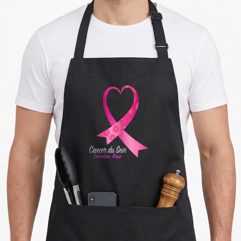 Tablier de cuisine noir avec un ruban rose stylisé en cœur et des fleurs, portant l'inscription Cancer du Sein Octobre Rose.