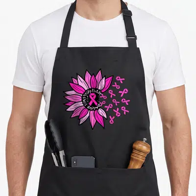 Tablier de cuisine noir avec une grande fleur rose, un ruban rose central et plusieurs petits rubans flottants sur le côté droit.