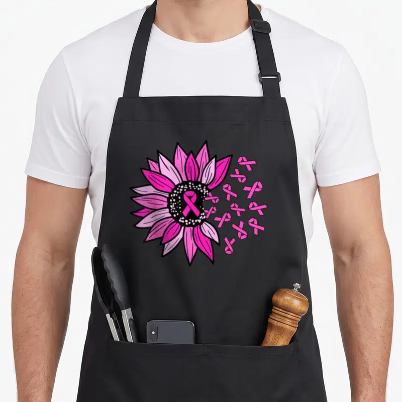 Tablier de cuisine noir avec une grande fleur rose, un ruban rose central et plusieurs petits rubans flottants sur le côté droit.