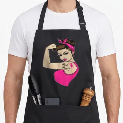 Tablier de cuisine noir avec illustration d'une femme aux cheveux chignon et bandeau rose, arborant un tatouage Pink Power sur le bras.