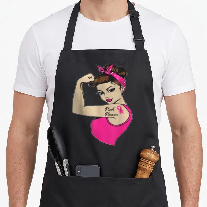 Tablier de cuisine noir avec illustration d'une femme aux cheveux chignon et bandeau rose, arborant un tatouage Pink Power sur le bras.