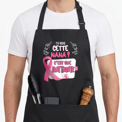 Tablier de cuisine noir avec un ruban rose et le texte Cette Nana Est Une Battante sur fond blanc.