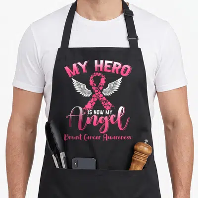 Tablier de cuisine noir avec un ruban rose en cœurs, des ailes blanches et le texte My Hero Is Now My Angel.