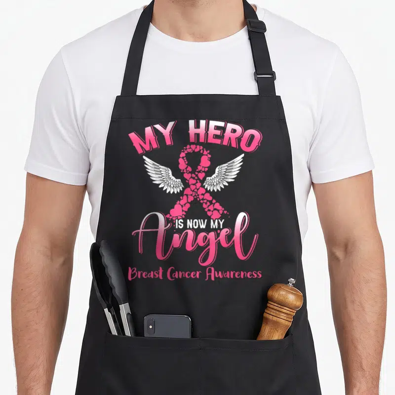 Tablier de cuisine noir avec un ruban rose en cœurs, des ailes blanches et le texte My Hero Is Now My Angel.