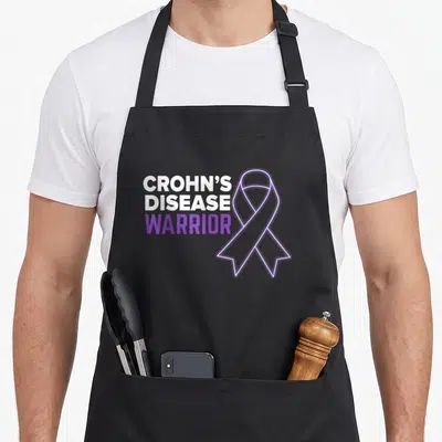 Tablier de cuisine noir avec inscription Crohn's Disease Warrior et un ruban violet stylisé.