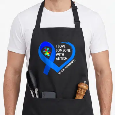 Tablier de cuisine noir avec un grand cœur bleu formé par un ruban et des pièces de puzzle colorées au centre.