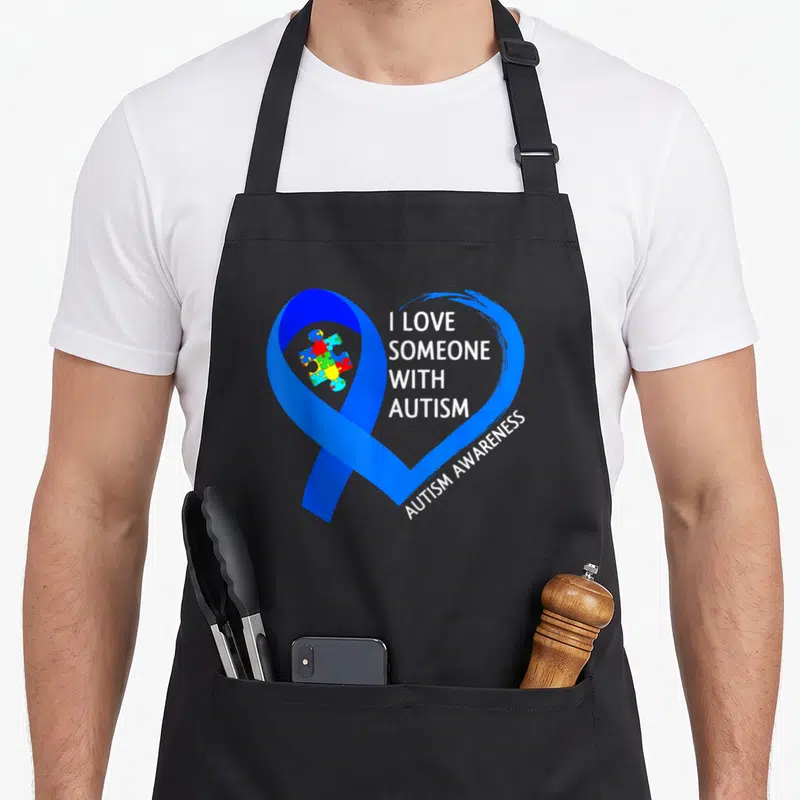 Tablier de cuisine noir avec un grand cœur bleu formé par un ruban et des pièces de puzzle colorées au centre.