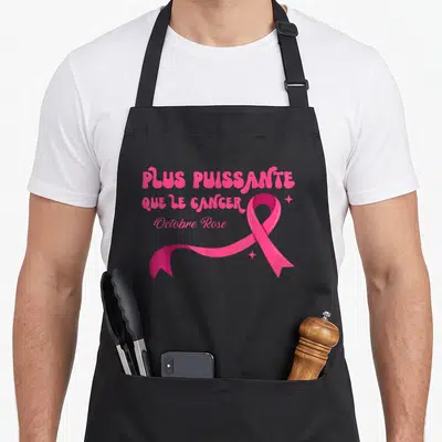 Tablier de cuisine noir avec l'inscription Plus Puissante Que Le Cancer et un grand ruban rose stylisé.