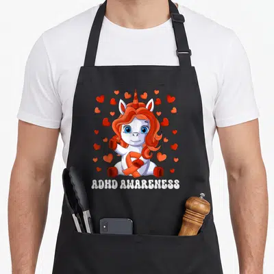 Tablier de cuisine noir avec licorne blanche, ruban orange et coeurs rouges autour du texte ADHD Awareness.