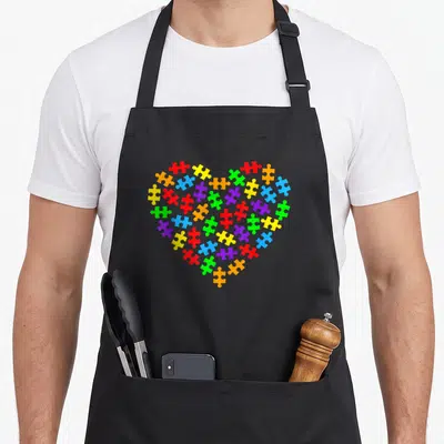 Tablier de cuisine noir avec un cœur formé par des pièces de puzzle colorées représentant l'autisme.