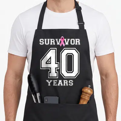 Tablier de cuisine noir avec linscription Survivor 40 Years et un ruban rose sur le devant.