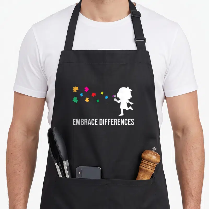 Tablier de cuisine noir avec silhouette enfant puzzle colorés et texte Embrace Differences en blanc.