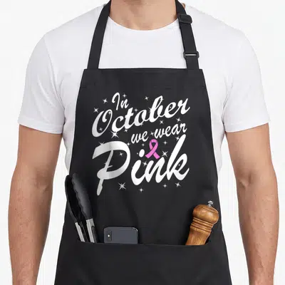Tablier de cuisine noir avec inscription blanche « In October we wear Pink » et un ruban rose entouré d'étoiles scintillantes.