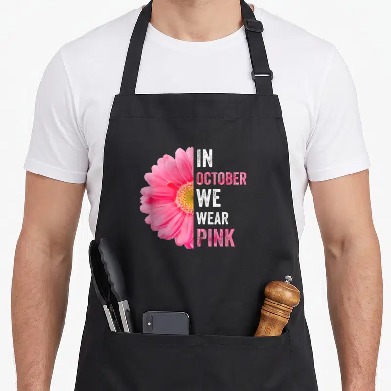 Tablier de cuisine noir avec une gerbe rose et le texte In October Nous Portons Rose imprimé en blanc et rose.