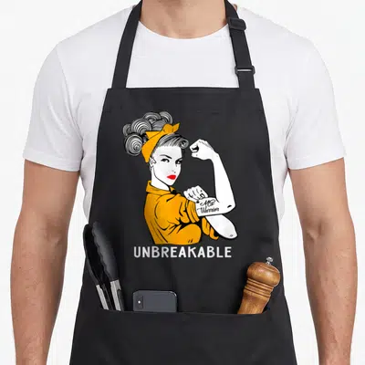 Tablier de cuisine noir avec illustration vintage d'une femme en orange et le texte UNBREAKABLE MS Warrior sur le bras.
