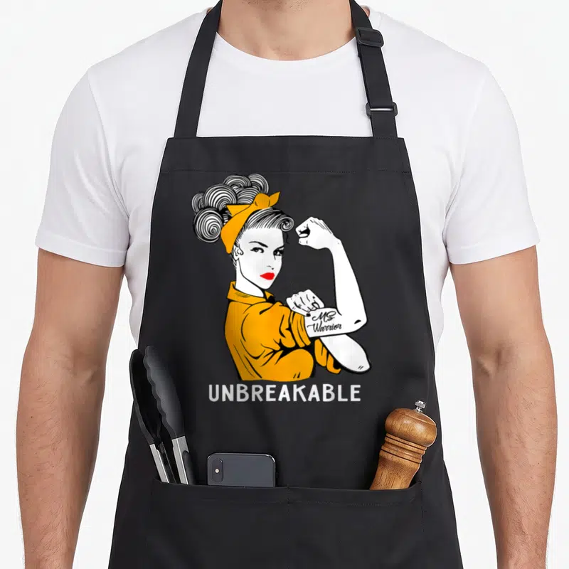 Tablier de cuisine noir avec illustration vintage d'une femme en orange et le texte UNBREAKABLE MS Warrior sur le bras.
