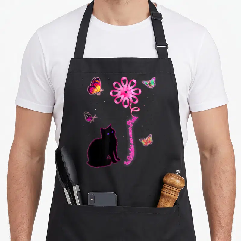 Tablier de cuisine noir avec un chat noir regardant un ruban rose en forme de fleur entouré de papillons multicolores.
