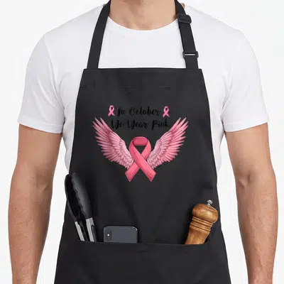 Tablier de cuisine noir avec un grand ruban rose orné d'ailes et du texte « In October Nous Portons Rose ».